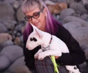 Woman with purple hair and glasses holding a white dog on rocky terrain.