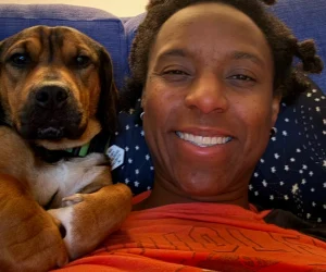 Happy woman with dog lying on a couch, smiling at camera, in a cozy home setting.