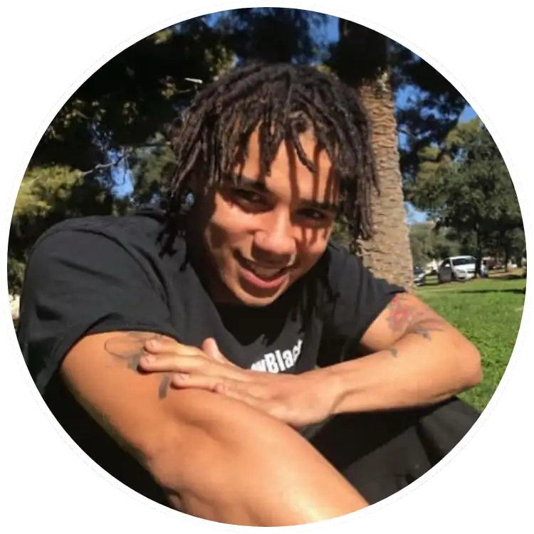 Young man with dreadlocks smiling outdoors in a park setting.